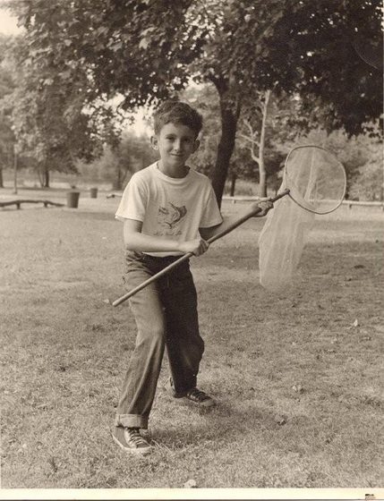 A sepia photograph of a boy standing on grass in a park, smiling at the camera and holding a long-handled butterfly net over his shoulder. He wears a t-shirt with a fish printed on it, rolled-up jeans and sneakers, with trees and a park bench behind him.